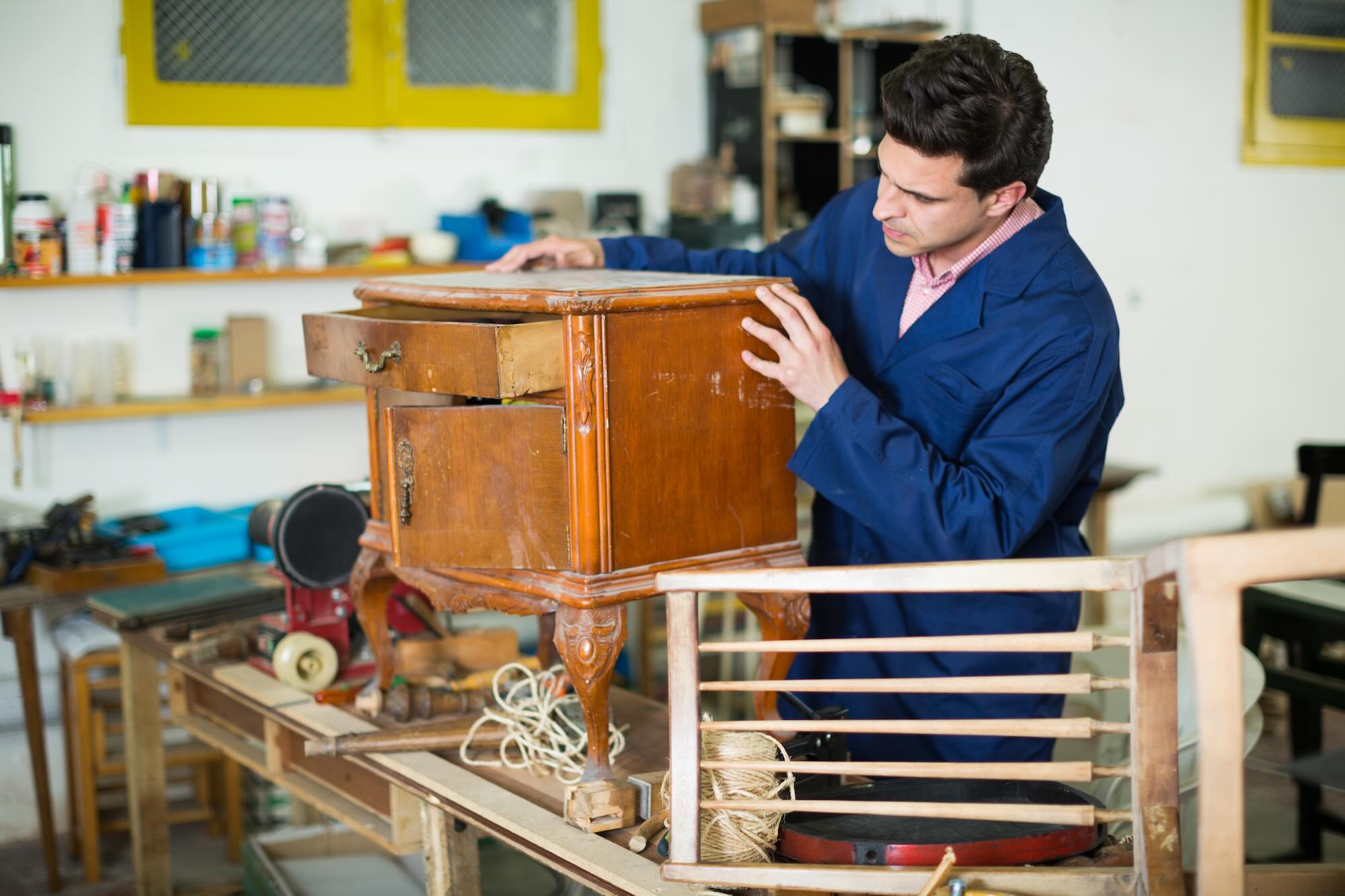 Man carpenter wearing blue overalls restoring wood furniture